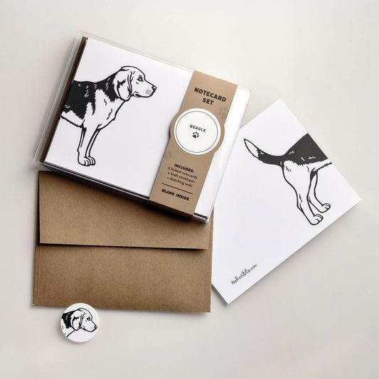 Beagle notecard set with clear box packaging and kraft belly band next to a folded card, kraft envelope, and matching round seal on a warm neutral background.
